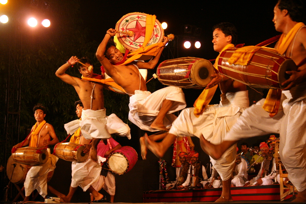 A photograph of musicians in traditional Manipuri wearing dhotis and dancing. Four musicians are carrying long drums and kicking in time, while two musicians playing colorfully decorated flat drums are in mid-air in what appears to be the first stage of a backflip.