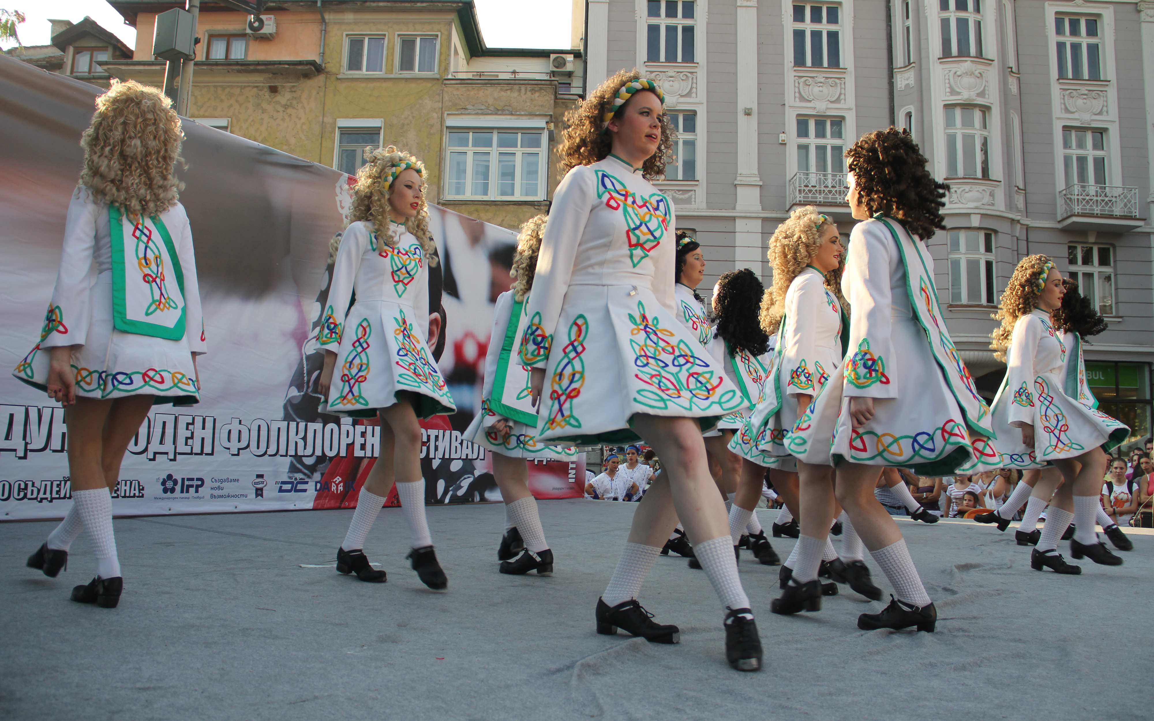 A photograph of a group of 10-15 women dressed in cultural Irish clothing, dancing in formation on an outdoor stage with townhouses visible in the background.