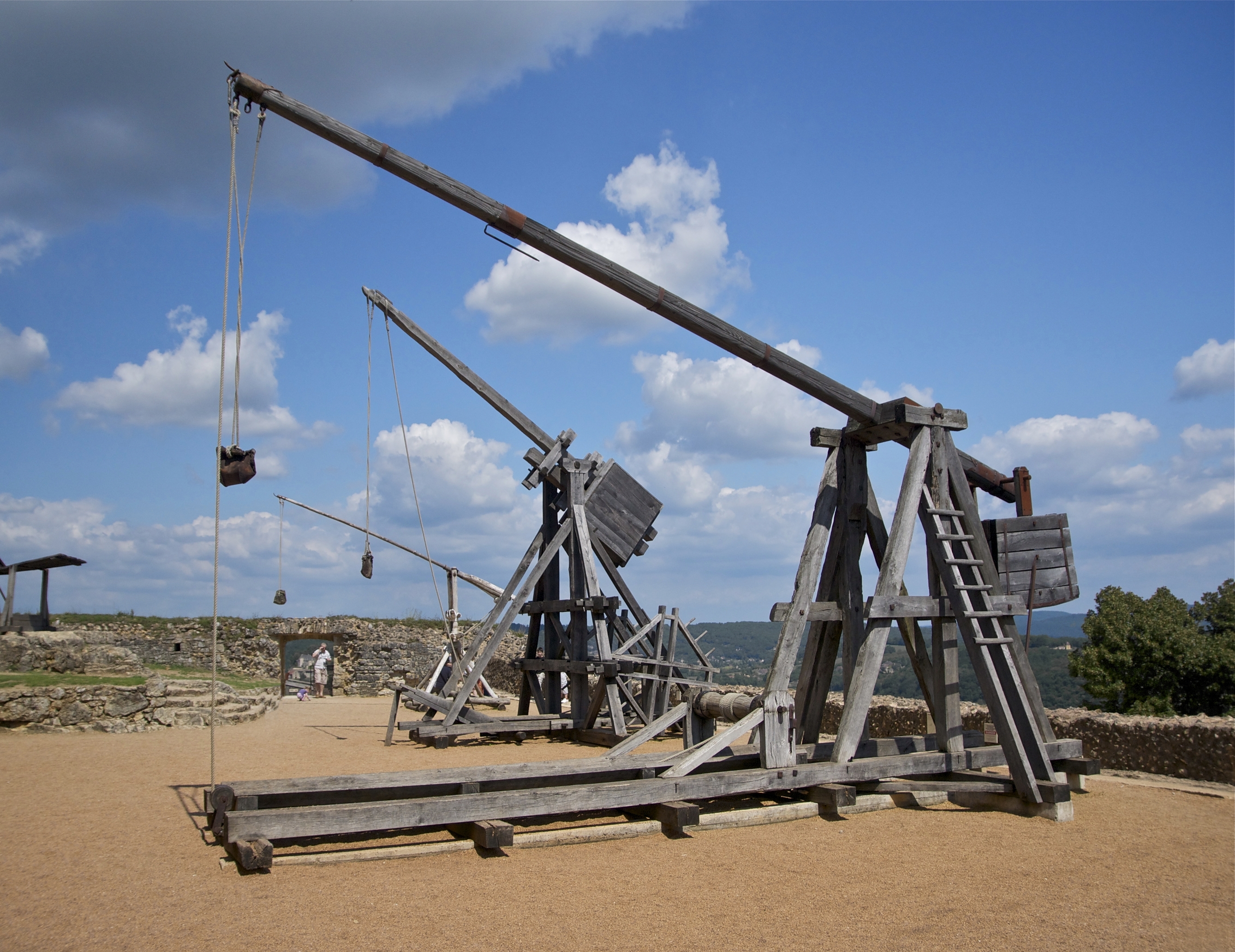 Three large wooden trebuchets sitting idly next to a low stone wall. Beyond the stone wall is a treetop and distant greenery, giving the impression that the foreground is atop a large castle.
