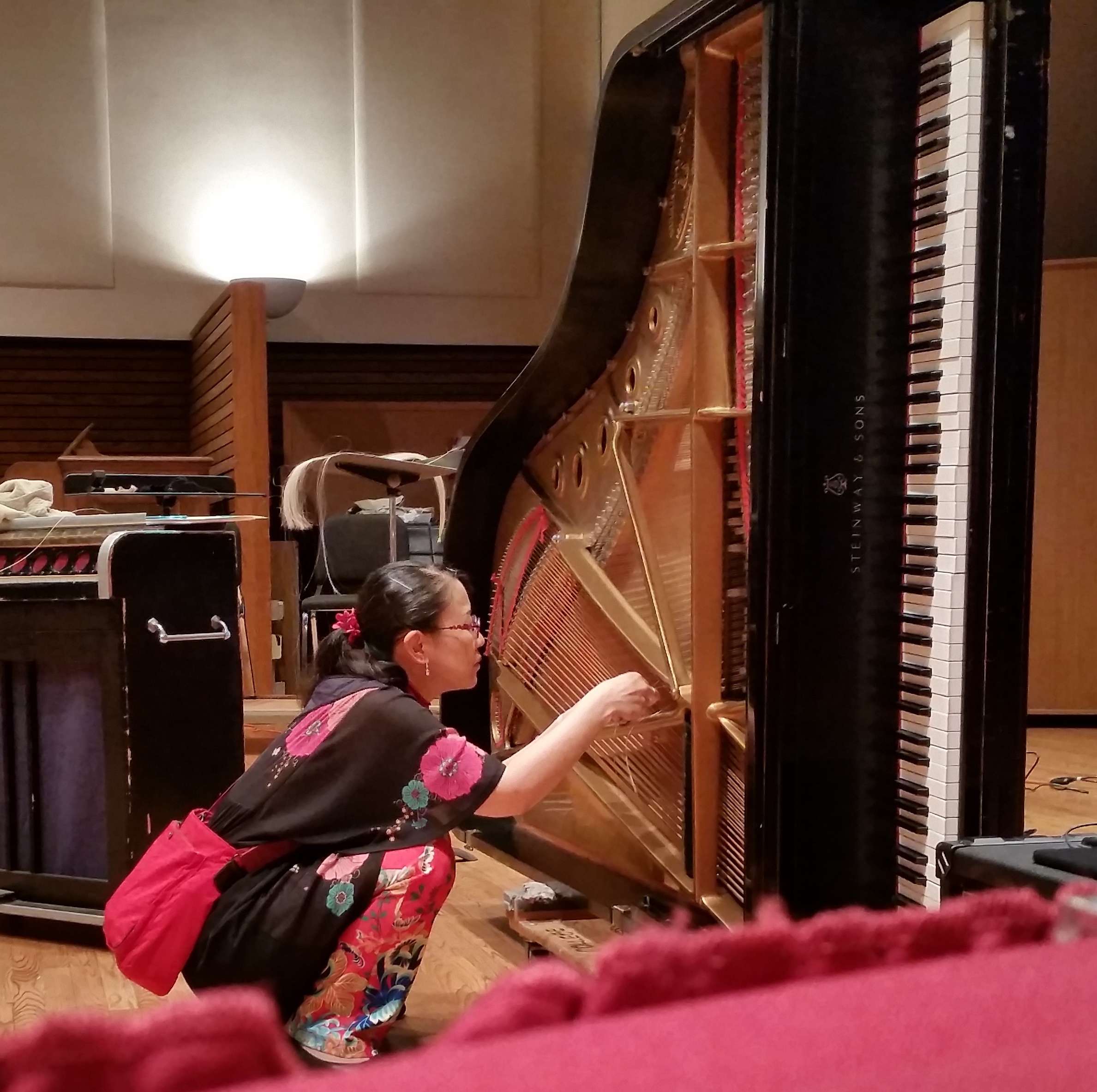 A woman crouches beside a grand piano which has been placed on its side on a stage. She is holding a fishing line which is woven between the strings of the instrument.