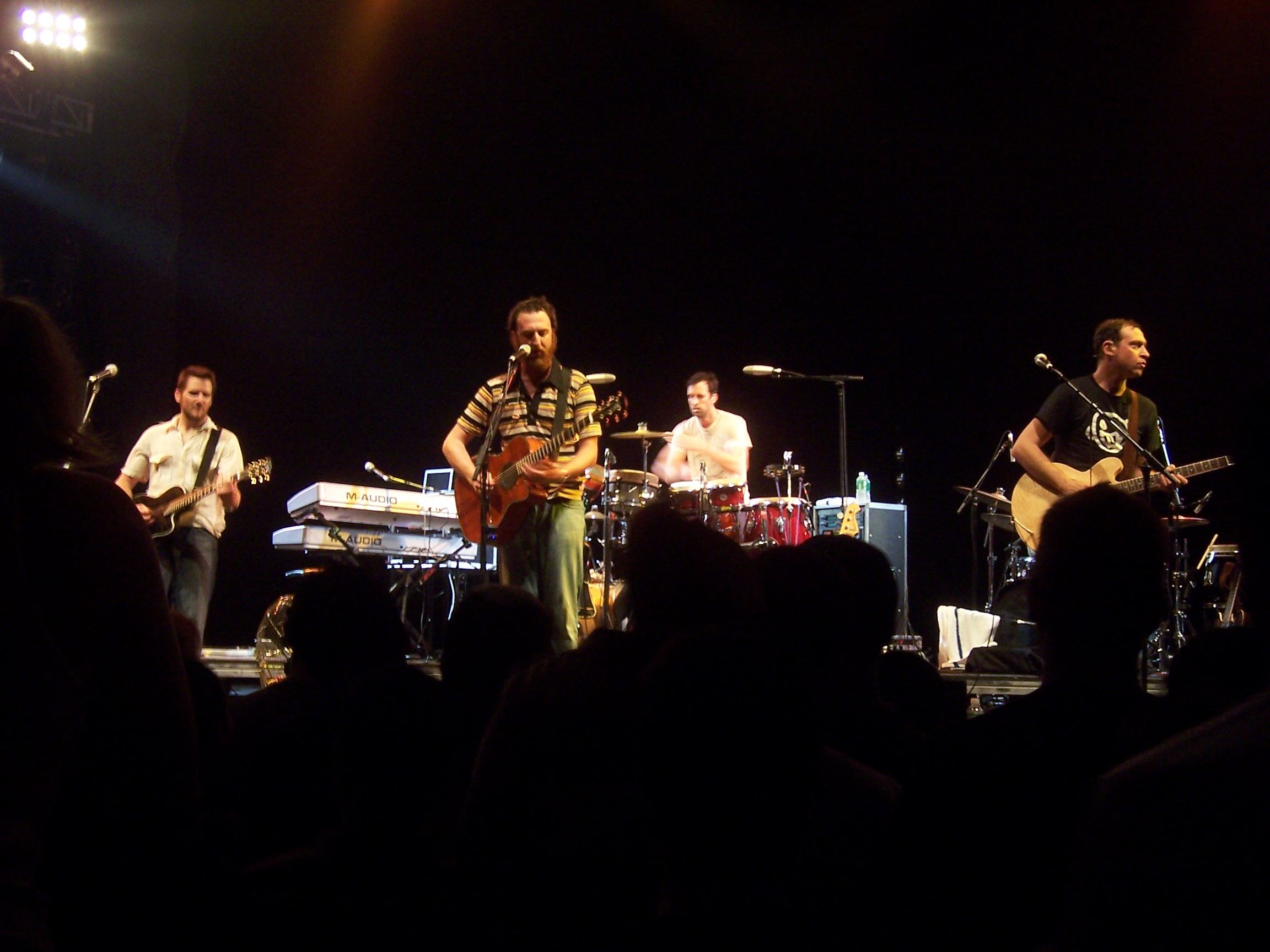 The four members of Guster perform on stage with a black backdrop behind them and the backs of audience members in the foreground. Three of the musicians are playing guitars and one is sitting at a drum set.