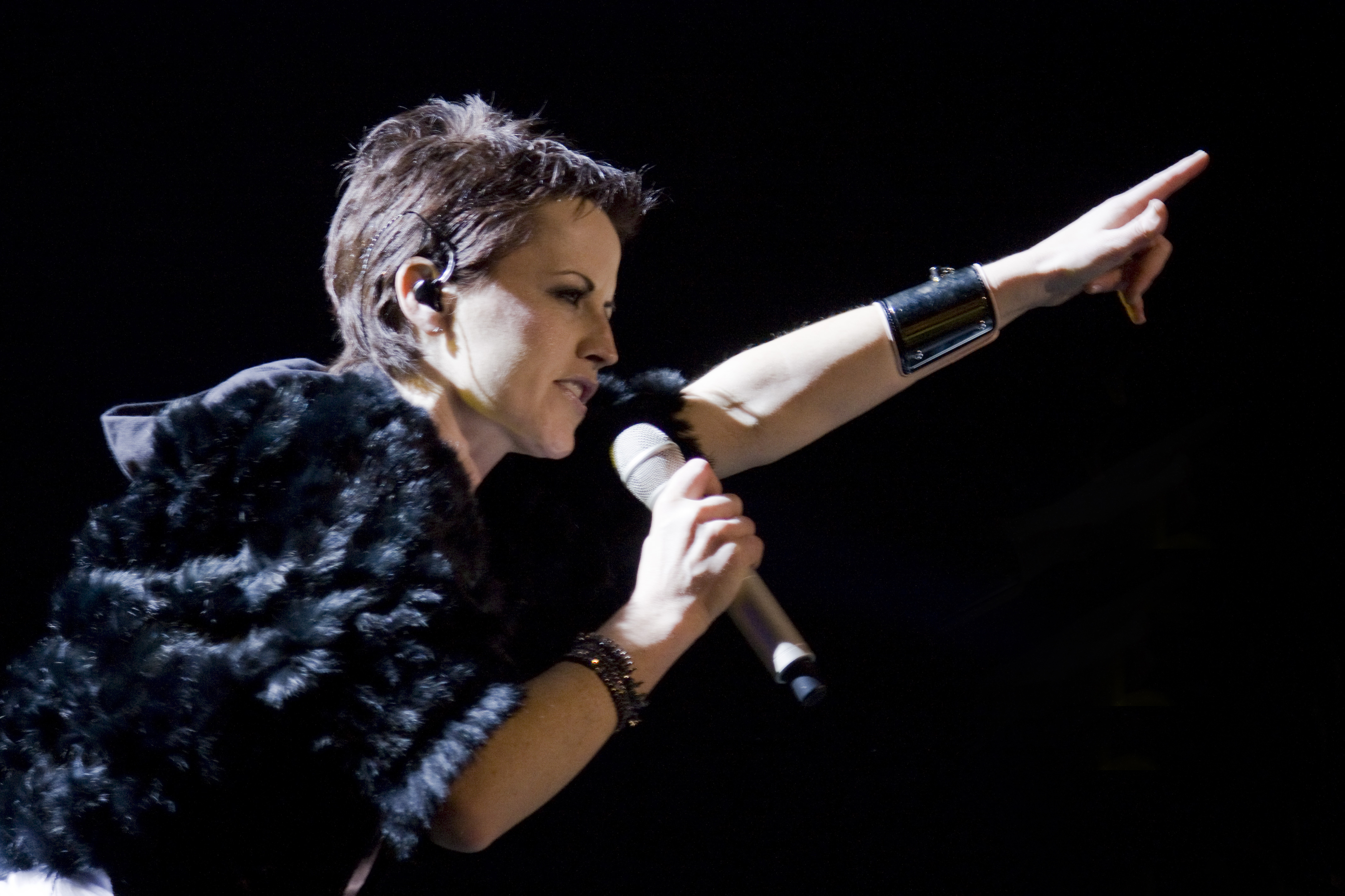 Dolores O'Riordan singing against a black backdrop, holding a microphone in her right hand and pointing outward to the audience with her left hand.