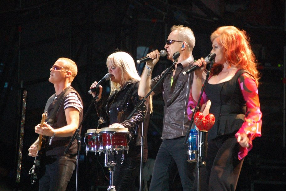 Members of the B-52s perform on stage. Wilson, Schneider and Pierson are singing into microphones, Wilson is also playing bongo drums, and Strickland is playing guitar.