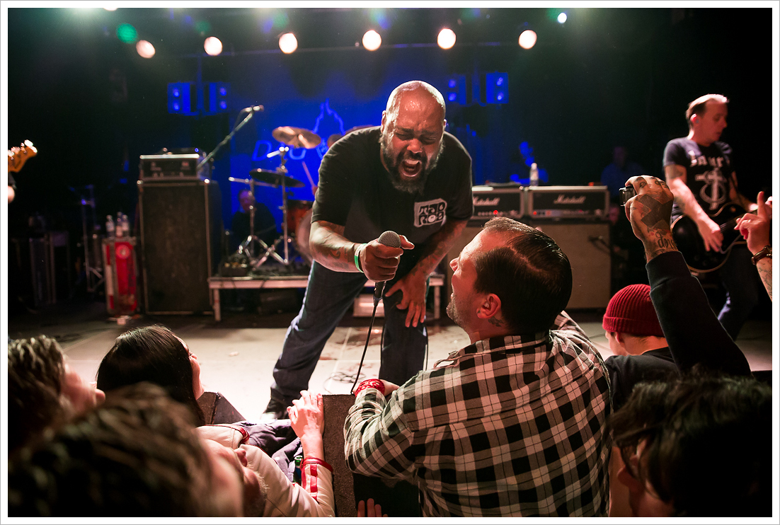 A photograph of the band Dag Nasty performing on stage, with lead singer Shawn Brown leaning into the front row of the audience and sharing his microphone with a fan singing along.