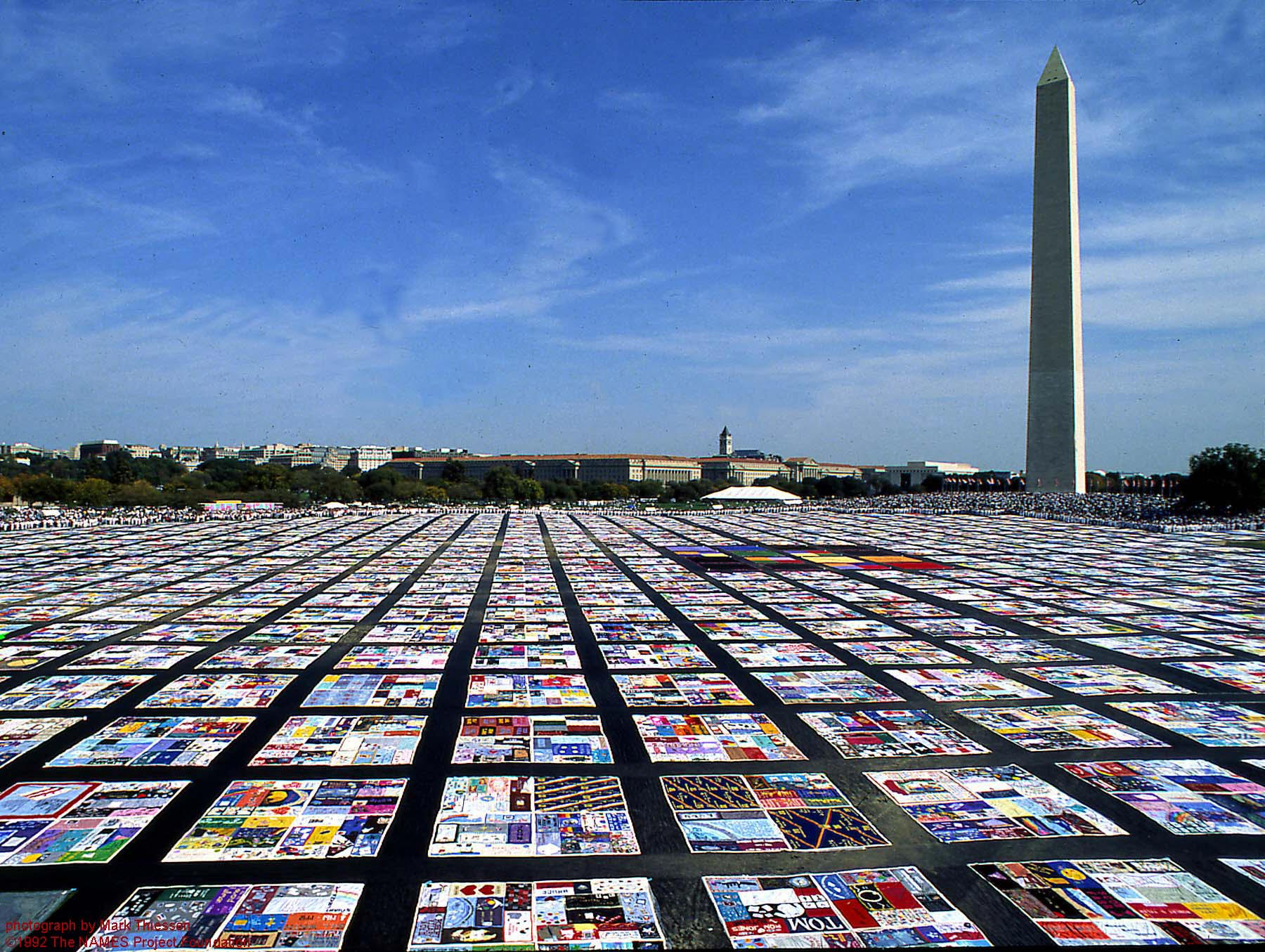 A photograph of the AIDS Quilt covering the National Mall in Washington, DC. Sections of the quilt are spaced equally to cover the entire Mall area, and the Washington Monument is seen in the background.