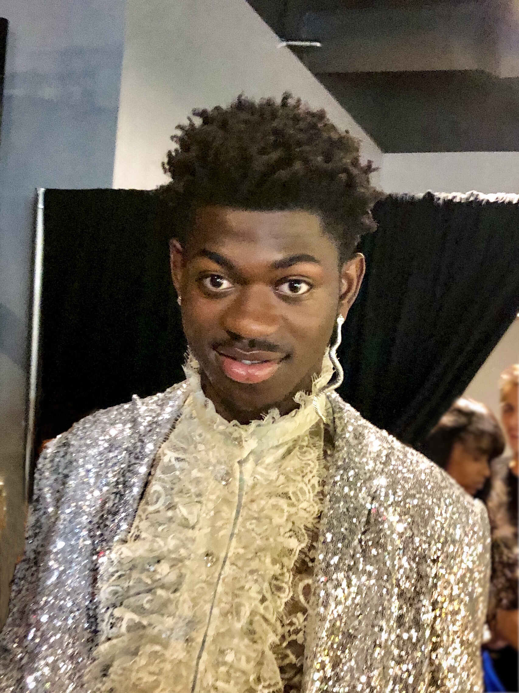 A photograph of Lil Nas X, wearing a lace shirt with ruffle and a silver sequined jacket.