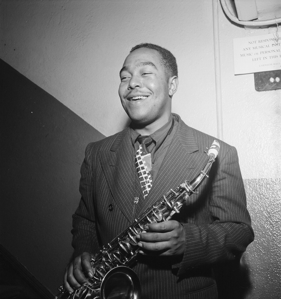 A black and white photograph of Charlie Parker in a backstage area, holding his saxophone and smiling as though he were in the middle of a friendly conversation with someone standing beside the photographer.