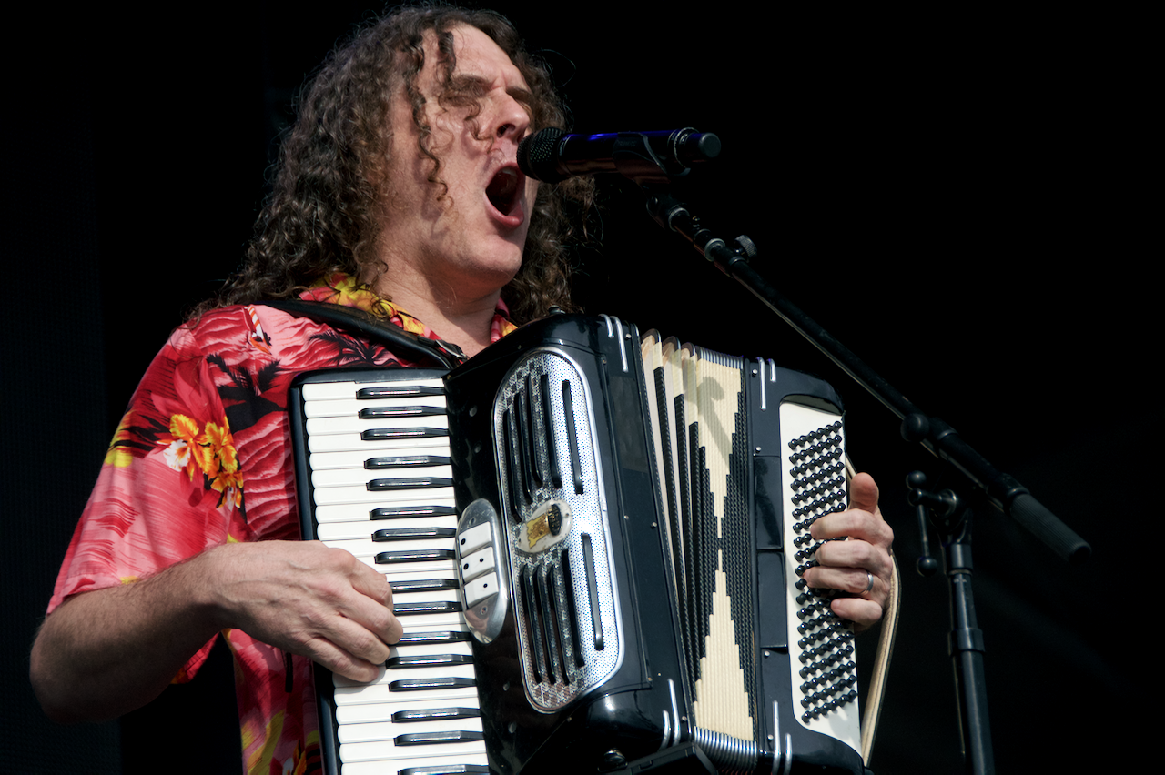 `Weird Al` Yankovic performing on stage, singing into a microphone while playing an accordion.