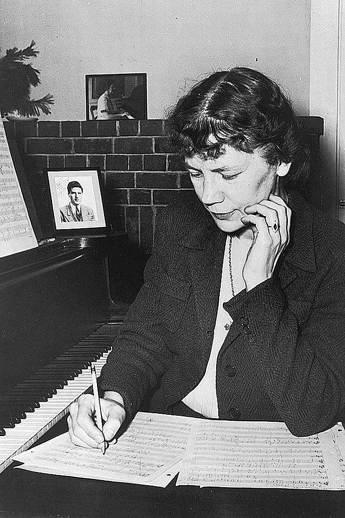 A black and white photograph of Louise Talma sitting at a piano and writing music on a large book of staff paper.