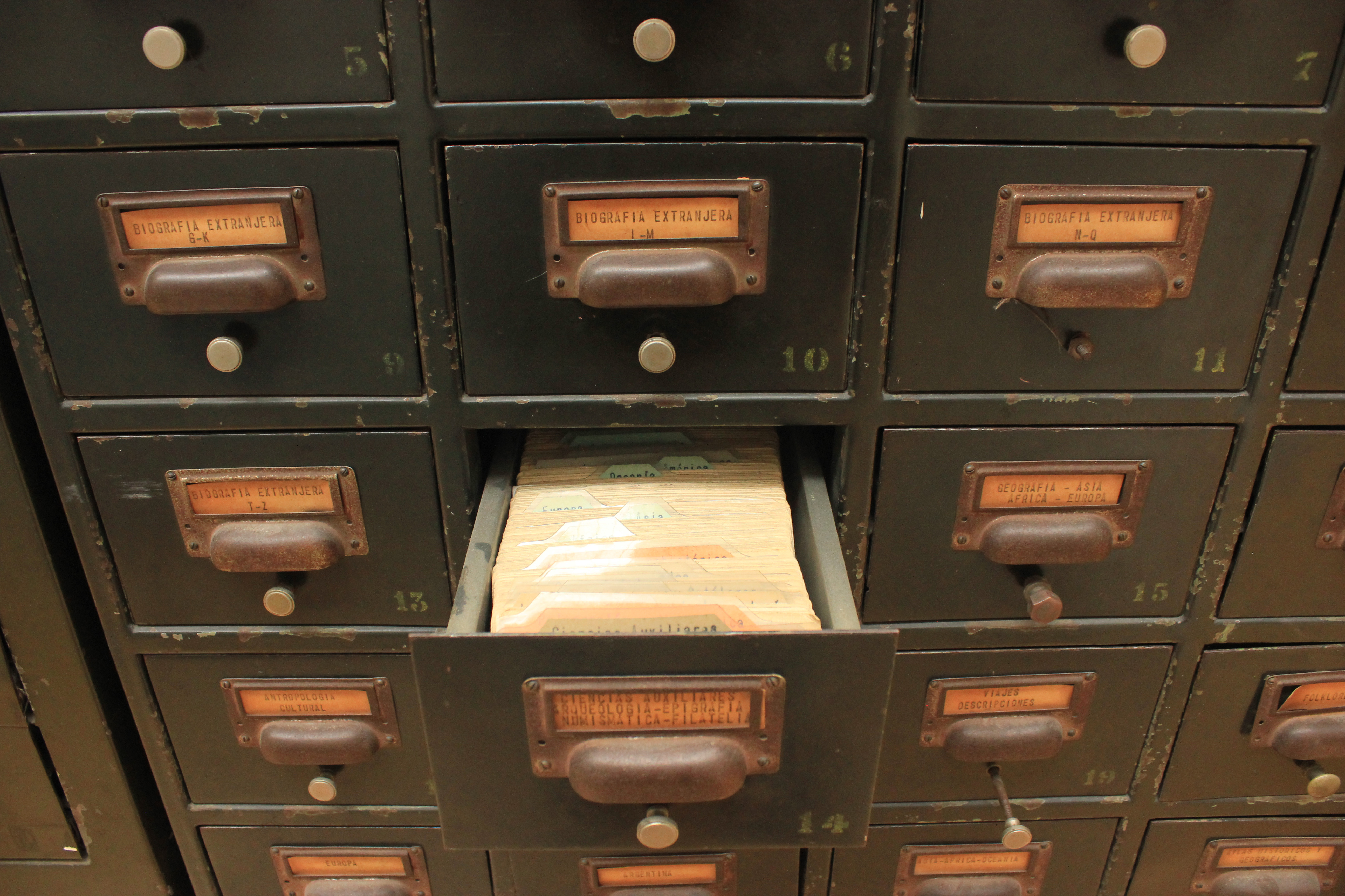 A closeup photograph of a physical library card catalog with Catalan labels on each drawer like `biografia extranjera` and `geografia - asia - africa - europa.` A drawer in the middle of the photograph labeled `sciencias auxiliares, arqueologia - epigrafia - numismatica - filatelia` is open, revealing a full stack of cards inside.