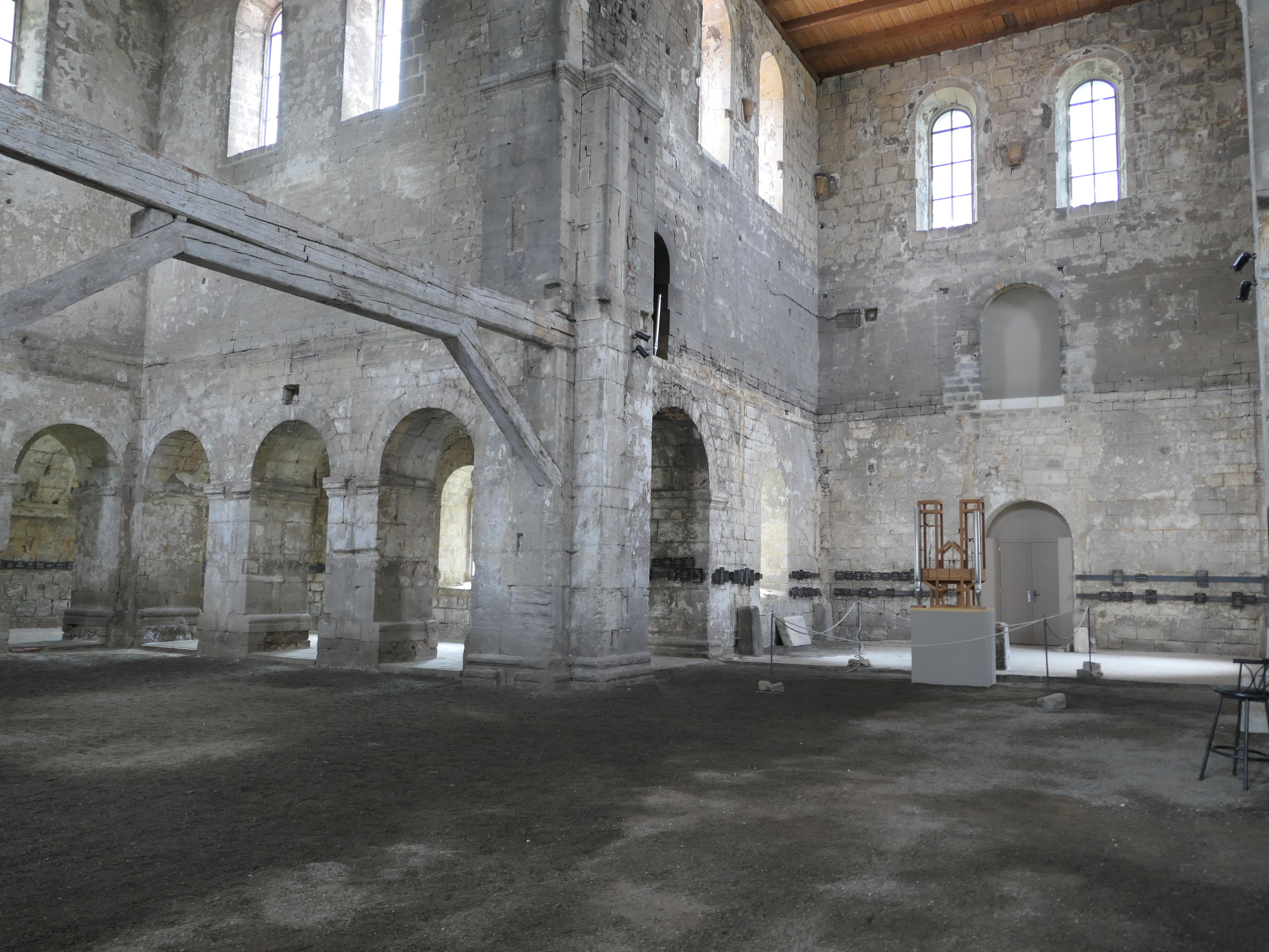 The undeveloped interior of an old stone church, showing old wooden beams, arched doorways and sunlit arched windows near the vaulted ceiling. In an alcove, cordoned off and dwarfed by the size of the room sits a special organ consisting of an open wooden structure and only three pipes.