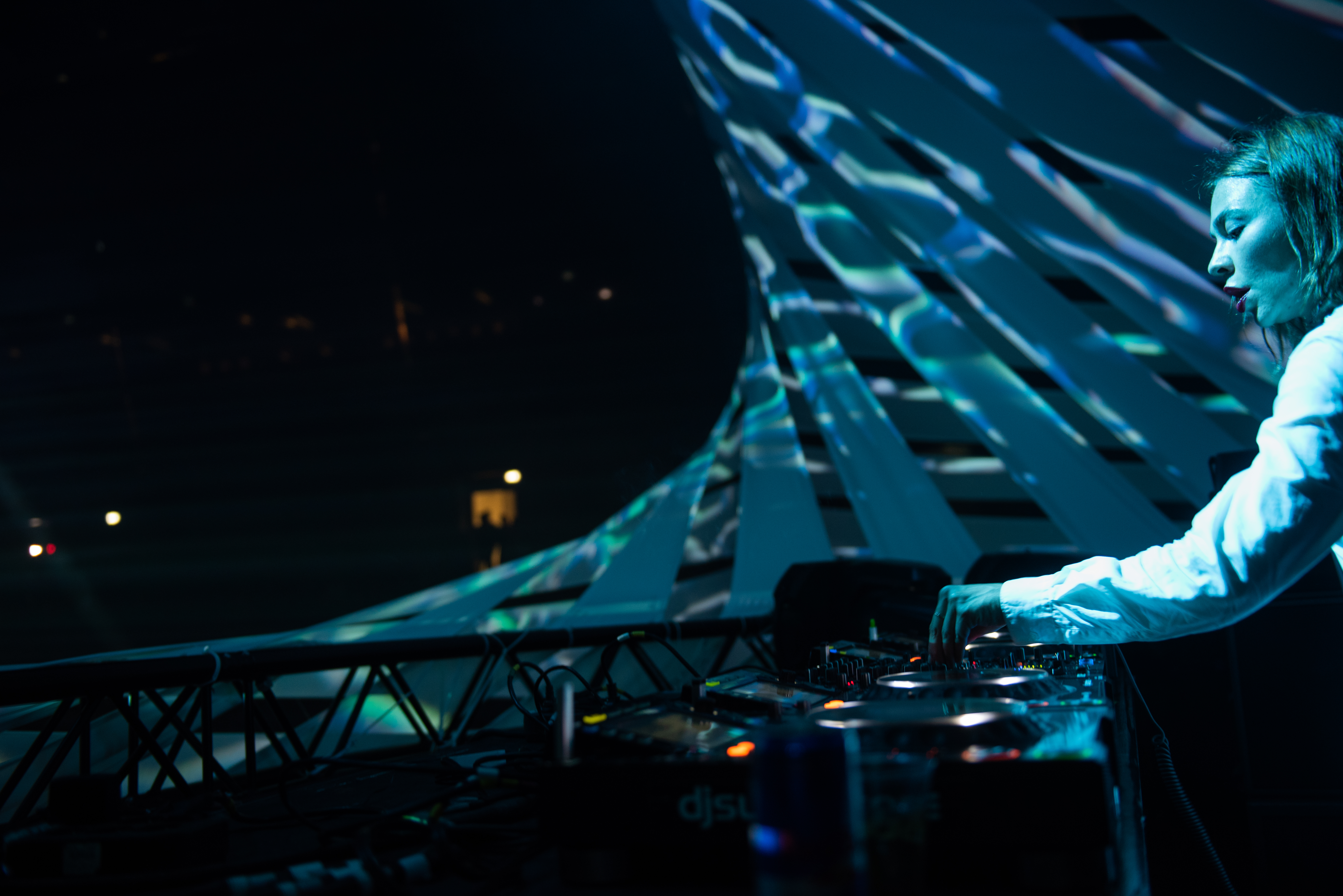 A photograph taken from stage left of DJ Nina Kravis performing onstage at her console under blue lights.